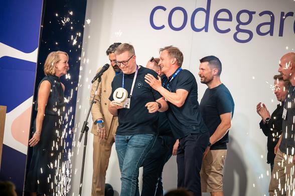 A joyful celebration on stage at Codegarden. A smiling team member holds a wooden Umbraco award while others gather around, clapping and cheering. Sparks fly at the edge of the stage, adding a festive touch. Everyone wears event lanyards, and the atmosphere is filled with excitement and camaraderie.