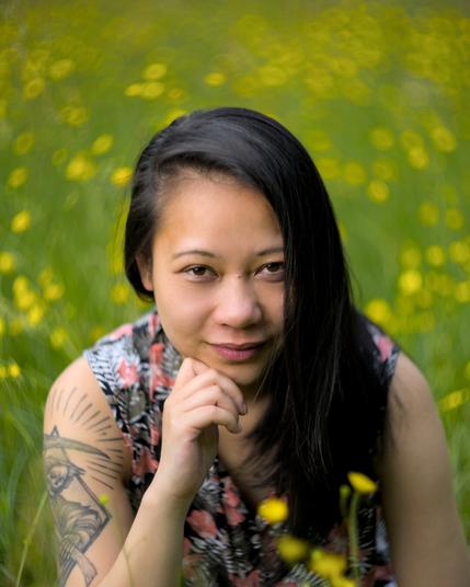 Eine Frau mit schwarzem Haar und tätowiertem Oberarm kniet in einer sommerlichen Blumenwiese. Der Hintergrund ist voller gelber Blüten und unscharf dank Vintage-Bokeh-Look. Sie blickt ruhig und direkt in die Kamera.