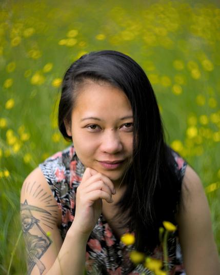 Eine Frau mit schwarzem Haar und tätowiertem Oberarm kniet in einer sommerlichen Blumenwiese. Der Hintergrund ist voller gelber Blüten und unscharf dank Vintage-Bokeh-Look. Sie blickt ruhig und direkt in die Kamera.