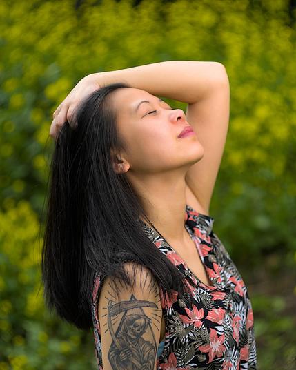 Eine Frau mit schwarzem Haar und tätowiertem Oberarm steht vor einer sommerlichen Blumenwiese. Der Hintergrund ist voller gelber Blüten und unscharf dank Vintage-Bokeh-Look. Sie blickt ruhig und direkt in die Kamera.