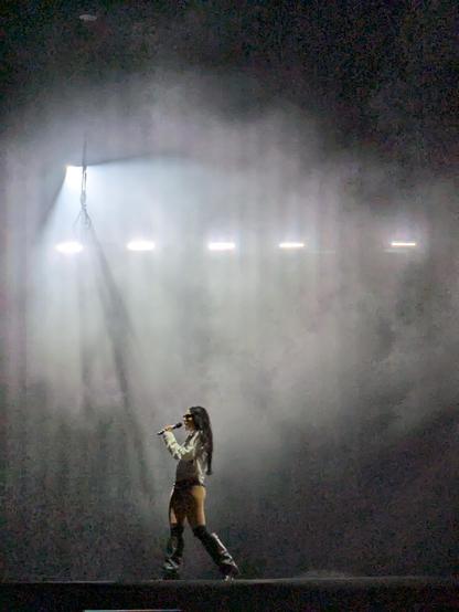 Charli XCX on stage at Primavera Sound Porto 2025. She is shown quite small against a backdrop of white lights striped by dry ice smoke. She has knee boots, a white top, and is apparently singing into the microphone, but probably just swearing