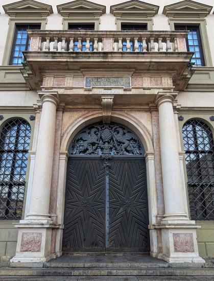 A grand, symmetrical close-up captures the ornate entrance of a historic building, Augsburg Town Hall, in a vertical composition. A large, dark wooden double door with a radiating star pattern on each panel is framed by two substantial fluted marble columns with ornate capitals. Above the door, an arched transom features intricate wrought-iron decoration, including what appears to be a pineapple and other motifs. The columns support a projecting balcony with a balustrade, and directly above the door, an inscription carved into stone reads "PUBLICO CONSILIO PUBLICO SALUTI". Two arched windows with decorative grilles flank the entrance. The building's light-coloured facade shows multiple upper windows with dark frames and simple balconies.