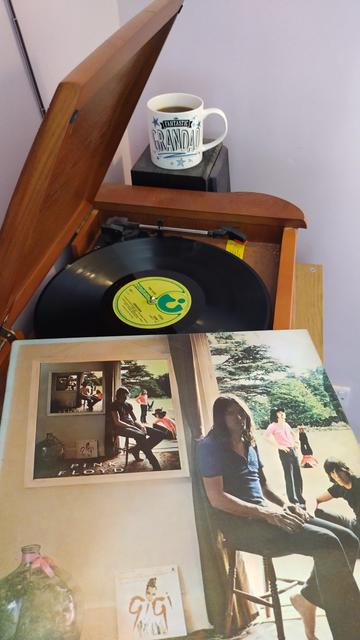 Record player with a 12" black vinyl record.
In the foreground is an album cover with the band seen on the right in various poses, from in an open doorway, out into a large garden.  On the wall on the left a picture hangs, showing the same scene, but with the band members swapping positions. Each picture repeats this effect, until the fourth pictures shows the album cover from their second album, A Saucerful Of Secrets.
Behind the player is a mug of coffee on a speaker.