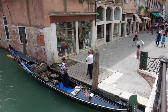 Looking down from a bridge over a Venetian canal at a gondolier, waiting in his moored gondola while a man and a woman discuss something. In the street behind them tourists are walking to and from.