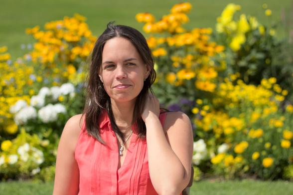 Une femme brune de face, cheveux longs, retiens ses cheveux de la main gauche, regard regardant sur le côté gauche, un sourire un poil crispé, sur un fond très fleuri à dominante jaune/orange/vert