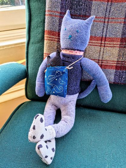A stuffed toy of an anthropomorphic blue cat wearing a pink and blue fuzzy shirt and white and black spotted socks. It is wearing a blue tote bag that has a small black cat tucked inside.