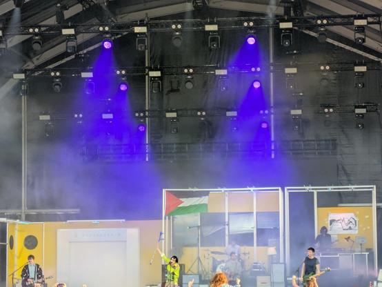 Badly framed photo of band on a festival stage - all stage, little band! Carolins Durante brought their own set, like an office in white  and yellow.
The band are spread across the stage. The singer is raising his crutch to the crowd. Blue lights beam down, and a Palestinian flag hangs central