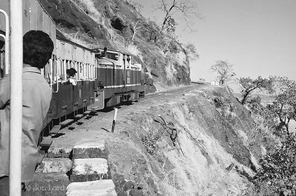 This Is An Older Black And White Street Photo In Landscape Format. Stretching From The Left Margin And Curving Round To The Right To The Centre Of The Image Is A Small Narrow Gauge Train. Closest To The Camera Are Two Small Carriages With A Man Hanging Onto The Side As Was Common On Indian Railways In The Past. Our Passenger Has Short Dark Hair And A Medium Grey Shirt. He Is Facing Away From The Camera Towards The Front Of The Train. The Carriages Have Open Windows Without Glass And A Number Of Passengers Can Be Seen Looking And Leaning Out Of The Windows. Ahead Of The Front Carriage Is A Diesel Locomotive With The Drivers Cab In the Centre, The Train's Driver Can Be Seen Leaning Out Of The Right Side Window. The Train Track Is Laid On A Narrow, Curving Terrace With A Steep Slope Above On The Left Side And An Equally Steep Slope Falling Away To The Right. The Track Is Curving To The Right Before Turning Sharply To The Left Where Is Disappears From Sight. There Is Little Vegetation To Be Seen, Just A Few Small Stunted And Bare Trees. 
The Location Is The Matheran Hill Railway That Runs 21km From Neral To Matheran, 90km East Of Mumbai, India (1990).