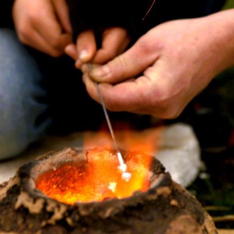 Kinderhände halten einen Stahldorn über die Feuerdüse eines Lehmofens. Eine Erwachsenenhand hilft mit.