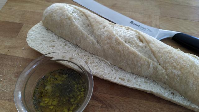 ein waagrecht halbiertes Baguette, eine Schüssel mit Öl, Knoblauch und Gewürzen sowie ein Brotmesser liegen auf einem Holzbrett