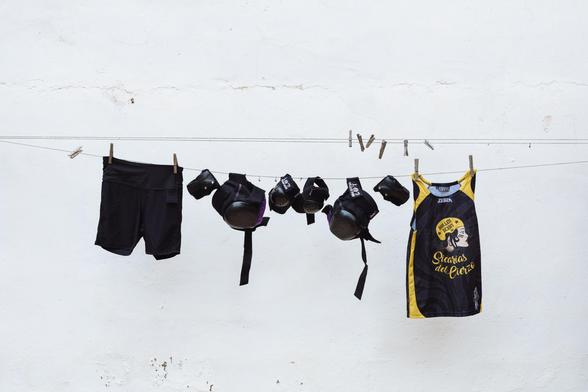 En un tendedero sobre fondo blanco, están colgadas la camiseta de las sicarias, unas mallas cortas negras y las muñequeras, las coderas y las rodilleras