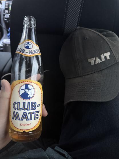 A shot of a 1 liter glass bottle of Club Mate soda held open in my left hand. A baseball hat embroidered with TAIT rests on my right knee