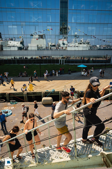 People climbing steps to board a warship that is itself reflected on the face of the building in the distance.