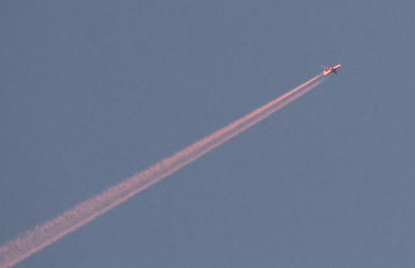Ein Flugzeug mit jeweils einem Düsentriebwerke an den Tragflächen, leuchtet rot - der Sonnenaufgang ist erst in 35 Minuten vorhergesagt. Des Weiteren sieht man am blauen Himmel keine Wolken  nur den Abgasstrahl des Flugzeuges, welcher ebenfalls in der Farbe rot erstrahlt. 
Matzework