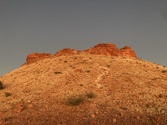 Bright dusk light on a steep orange rocky hill with a path winding up to redder orange rock outcrops at the top. The sky is an even grey.