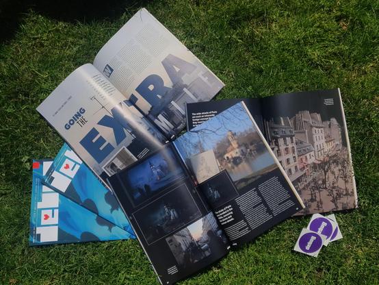 5 copies of Debug Magazine, with 3 spread open showing a article titled 'Going The Extra Mile' laying outside on grass