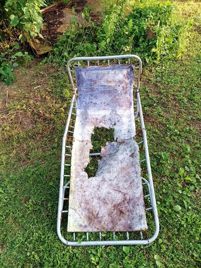 A completely knackered folding sun lounger. The frame is broken, hinges rusty, fabric decayed with a huge hole torn through the middle. It's standing outside in mowed grass.
