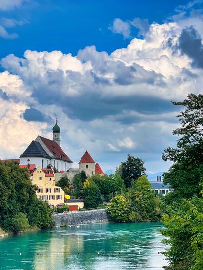 Under a deep blue, but clearly cloudy sky, a church on a hill dominates the photo on the left. Other historic buildings can be seen next to and below the church, framed by trees. The buildings end in the lower part of the image with a river wall. Below flows a greenish river, the Lech. Several swans can be seen. On the right, trees protrude into the picture, framing the summery scene.