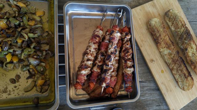 Die fertig gegrillten marinierten Hähnchenspieße mit speckumwickeltem Grillkäse liegen wie auch die Antipasti in je einer Edelstahlform, daneben das Knoblauchbaguette auf einem Holzbrett