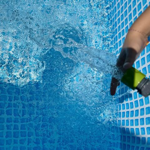 Schatten auf dem PoolBoden in Form eines Kopfes, Schlauch der Wasser in den Pool spritzt, eine kleine Hand vor dem Wasserstrahl, die Wasseroberfläche verwirbelt