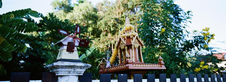Panoramic photo of a small wooden windmill on a white stone pedestal next to an intricately decorated gold and brown shrine structure . The background features lush green foliage and a clear blue sky.