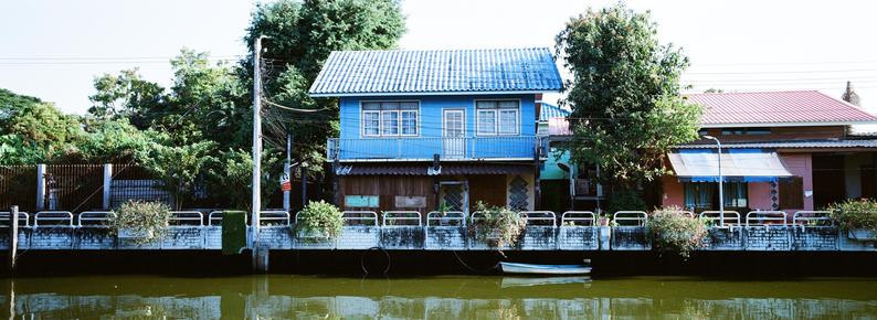 Panoramic photo of a blue two-story house with a tiled roof and front porch, flanked by trees on both sides. To the right is a beige single-story building with a red roof and an awning. A canal runs in the foreground, with a small blue boat tethered to the cement barrier. Lush greenery surrounds the area, and a metal railing with flower pots lines the edge of the canal.