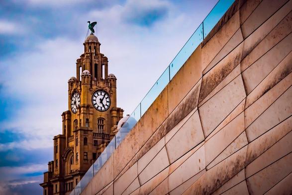 Clock tower with a bird statue on top, featuring large black and white clock faces. The background shows a partly cloudy sky. In the foreground, a modern beige wall with geometric patterns and a glass barrier on top runs diagonally across the image.