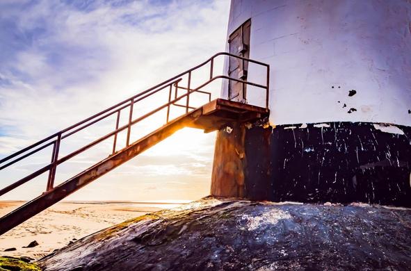 Rusty metal staircase attached to the side of a weathered lighthouse with peeling black and white paint. Sunlight shines from behind, casting a glow on the structure. Sandy beach and cloudy sky in the background.
