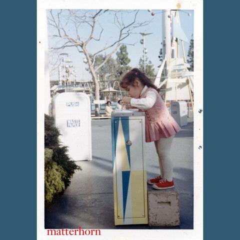A young girl stands on a concrete block, reaching into a colorful, vertically striped box in an amusement park setting. She is wearing a pink dress and white leggings, with red shoes. Background features amusement park attractions and trees.