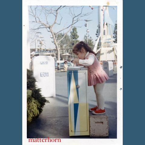 A young girl stands on a concrete block, reaching into a colorful, vertically striped box in an amusement park setting. She is wearing a pink dress and white leggings, with red shoes. Background features amusement park attractions and trees.