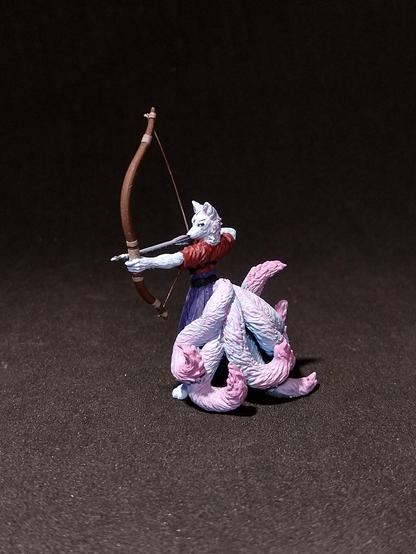 Das Bild zeigt eine detaillierte Miniaturfigur eines Wesens, das wie ein Wolf aussieht, das einen Bogen spannt. Das Wesen hat eine weiße Felle und trägt ein rotes Oberteil mit einem schwarzen Gürtel und einen langen, violetten Rock. Der Bogen ist aus Holz und hat eine geschwungene Form, die von den Händen des Wesens gehalten wird. Der Schaft des Bogens ist dünn und braun, und die Sehne ist sichtbar. Der Hintergrund ist dunkel, was die Figur hervorhebt. Die Figur steht auf einem flachen, dunklen Untergrund, der die Details der Figur betont.