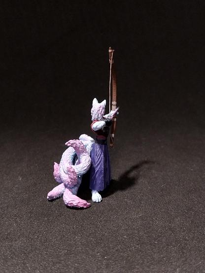 Das Bild zeigt eine detaillierte Miniaturfigur eines Wesens, das wie ein Wolf aussieht, das einen Bogen spannt. Das Wesen hat eine weiße Felle und trägt ein rotes Oberteil mit einem schwarzen Gürtel und einen langen, violetten Rock. Der Bogen ist aus Holz und hat eine geschwungene Form, die von den Händen des Wesens gehalten wird. Der Schaft des Bogens ist dünn und braun, und die Sehne ist sichtbar. Der Hintergrund ist dunkel, was die Figur hervorhebt. Die Figur steht auf einem flachen, dunklen Untergrund, der die Details der Figur betont.