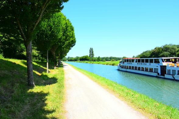 A white riverboat named “Santa Monika” sails along a quiet canal next to a tree-lined gravel cycle path under a clear blue sky.

Ein weißes Flussschiff namens „Santa Monika“ fährt auf einem ruhigen Kanal neben einem von Bäumen gesäumten Kiesradweg unter einem klaren blauen Himmel.