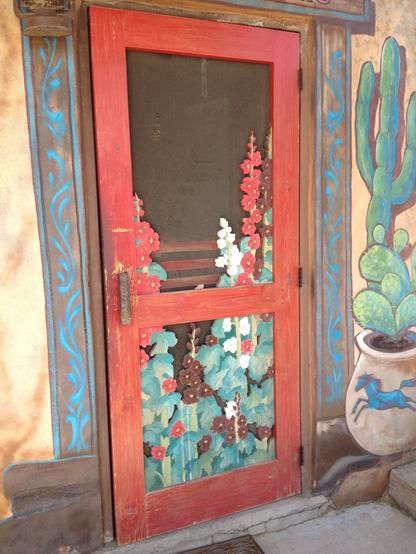 A weathered screen door with trompe l'oeil hollyhocks on it. To the right of the screen door is a trompe l'oeil planter on the wall with painted cactus emerging from the top