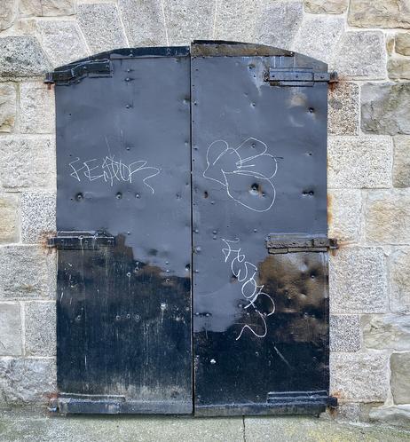 Old black painted metal doors set into a stone wall. Various tags on door.