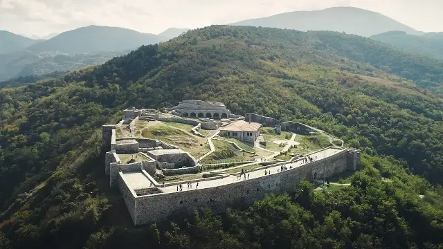 The image shows an aerial view of an ancient fort or castle situated on a hillside. The structure features stone walls and several buildings, surrounded by greenery and hills in the background. The landscape appears to be mountainous, with trees and diverse terrain surrounding the fortress. Visitors can be seen walking around the site, indicating it's a popular location for exploration or tourism.

Image Credits: Wikimedia / WeBalkans EU / CC BY 2.0