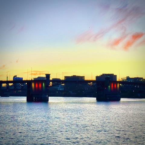 The Morrison Bridge lights up in red, yellow, and green colors over the Willamette River just before sunrise under a yellow, blue, and pink-cloudy sky.