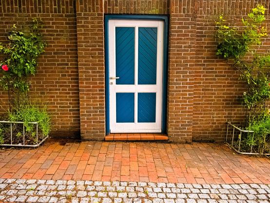 A blue door with a white frame and a white "cross" in the middle is set in a red brick wall. To the right and left of the door are rose bushes with a few pink roses.