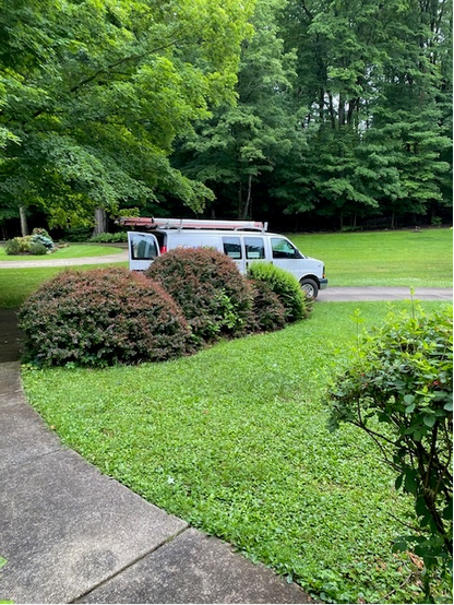Color image of an HVAC company van parked in the driveway along with two more, overhauling a residential HVAC system on the last day of spring. Image shows a white commercial work van parked in a driveway, ladders on top, door open, beyond a yard and hedges and a front walk.