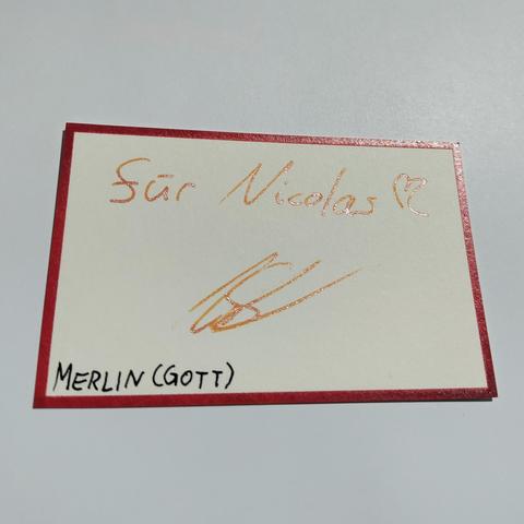 A paper card with a red border, with the Text "für Nicolas" and a signatute in shimmering orange ink, subtitled "Merlin (Gott)"