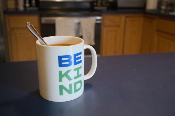 A mug of tea on a counter in a kitchen. There’s a spoon sitting in the mug and the mug reads BE KIND.