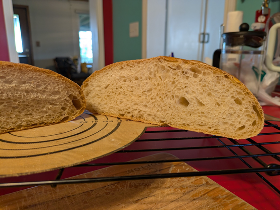 Halves of a boule bread loaf on a black wire cooling rack, positioned above a wooden cutting board. The bread has a good, open crumb structure with irregular holes throughout. The crust is golden brown. A bit of a home kitchen can be seen in the background."