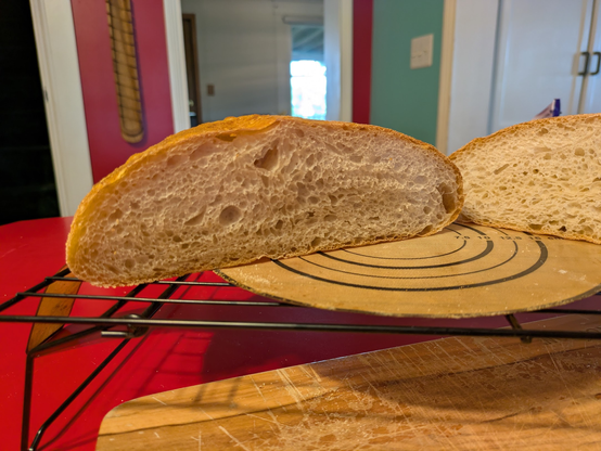 "Close-up of a sliced boule bread loaf on a black wire cooling rack, positioned above a wooden cutting board. The bread has a good, open crumb structure with irregular holes throughout. The crust is golden brown."