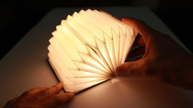 A half opened book lamp on a table unfolding it's bellows. It lights up bright tinting the table in warm white light. The book is opened by two hands with are also illuminated by the book lamp.