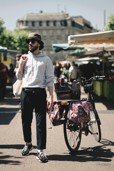 A bearded man wearing a bucket hat, sunglasses, and a light hoodie stands beside a bicycle with colorful floral saddle bags at an outdoor market. He carries a tote bag over his shoulder, with a relaxed posture, surrounded by people and market stalls under canopies on a sunny day.