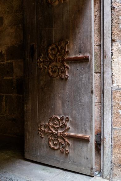 Eine offene Tür zu einem alten Gebäude. Die Tür besteht aus massiven Holzbrettern und hat schöne schmiedeeiserne Verzierungen, die die Bretter zusammenhalten. Der Fokus des Fotos liegt auf den Dekorationen.

An open door into an old building, the door is made of solid wooden boards and has beautiful wrought iron decorations that keep the boards together. The photo focuses on the decorations.