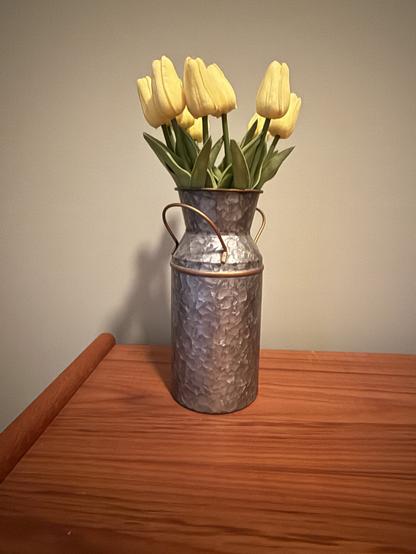 A rustic metal jug holds vibrant yellow tulips with green leaves, set on a wooden table against a plain background, evoking a cheerful and cozy mood.