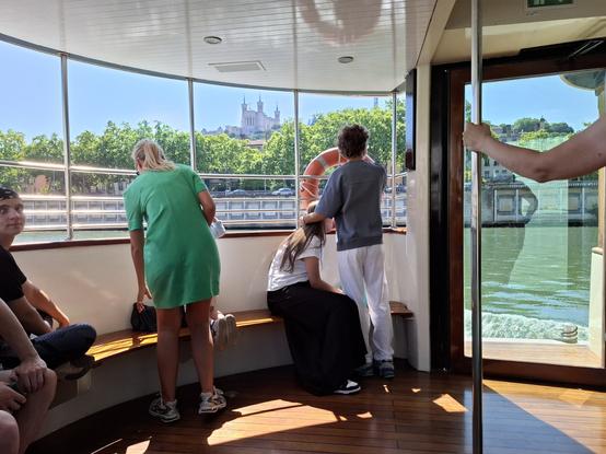 Arrière du bateau, des gens sont installés sur les bancs, en arrière plan la basilique de Fourvière