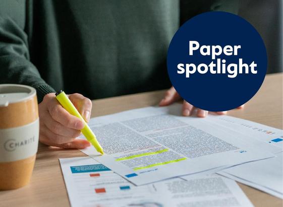 A person marks text passages on a scientific article with a yellow highlighter. There are several printed pages on the table, with a cup labeled “Charité” next to it. At the top right is a dark blue circle with the white text “Paper spotlight”.