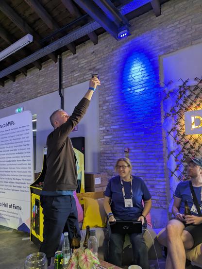 A tall man holds a mobile phone above his head while onlookers observe.
